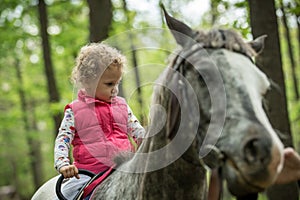 Girl enjoying horseback riding in the woods, young pretty girl with blond curly hair on a horse with backlit leaves