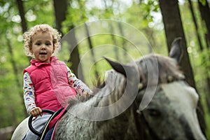 Girl enjoying horseback riding in the woods, young pretty girl with blond curly hair on a horse with backlit leaves