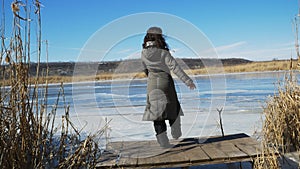 The girl enjoys the peace and beauty of nature, walking on a wooden pier on the bank of a frozen river