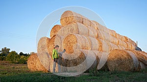 A girl examines large rolls of bales of hay in a field at sunset. The rolls are stacked in a pyramid shape