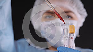 Girl examines a test tube with biomaterial. Young woman in protective clothing. On the face of a protective medical mask. A drop o