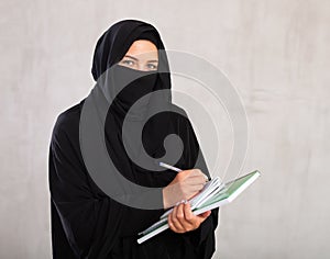 girl with face covered by black burka holds several notebooks and writes important note in diary