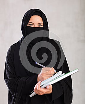 girl with face covered by black burka holds several notebooks and writes important note in diary