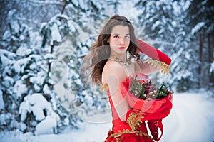 A girl in a fairy-tale image of a queen poses in a snow-covered winter forest. Long red dress, bouquet of fruits, red apples.