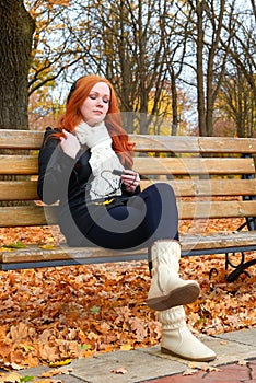 Girl in fall season listen music on audio player with headphones, sit on bench in city park, yellow trees and fallen leaves