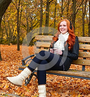 Girl in fall season listen music on audio player with headphones, sit on bench in city park, yellow trees and fallen leaves