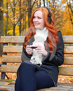 Girl in fall season listen music on audio player with headphones, sit on bench in city park, yellow trees and fallen leaves