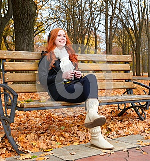 Girl in fall season listen music on audio player with headphones, sit on bench in city park, yellow trees and fallen leaves