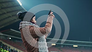 Girl fan taking picture of football match. Female spectator watching sport game at open air illuminated with stadium