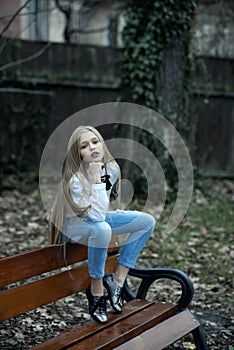 Girl in fashionable jeans sit on bench, fashion. Small child with long blond hair, hairstyle outdoor beauty. Baby beauty