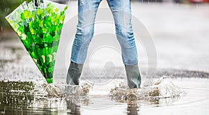 Girl feet in rubber boots with umbrella under rain in puddle