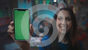 A girl in a festive hat shows a smartphone with a green screen, smiles. Close up of green screen smartphone on a blurred