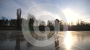 Girl in figure skates is skating on ice surface at sunset time. Young woman training alone at frozen river or lake. Lady