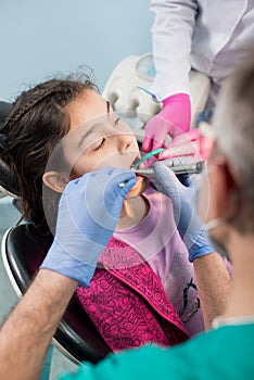 Girl with on the first dental visit. Senior pediatric dentist with nurse treating patient teeth at the dental office