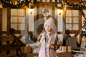 A girl of five or six years in a pink knitted hat with a pompom in a pink fur vest stands against