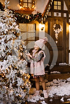 A girl of five or six years in winter pink clothes and ugg boots stands near the Christmas tree and decorates it with balls on the