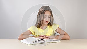 Girl flips through the pages of a book, and reads a book sitting at a table on a white background. The girl learns to