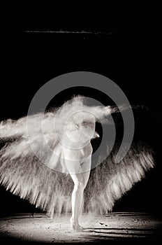 The girl with the flour on the body stretches the arms up with thrown flour on black background black and white image