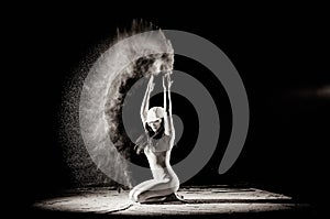 The girl with the flour on the body stretches the arms up with thrown flour on black background black and white image