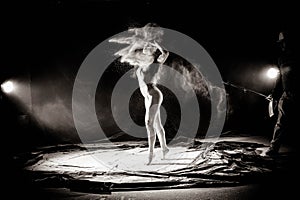 The girl with the flour on the body stretches the arms up with thrown flour on black background black and white image