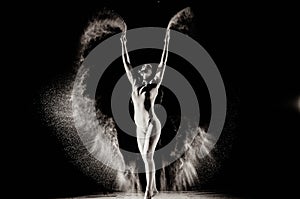 The girl with the flour on the body stretches the arms up with thrown flour on black background black and white image