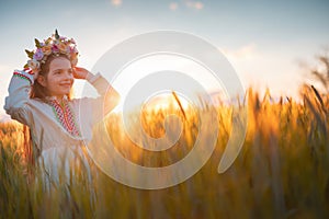 Girl with flower chaplet, ethnic folklore dress with traditional Bulgarian embroidery during sunset in wheat field