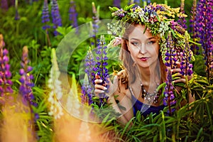 A girl with a flower wreath on her head sits at sunset in a field with purple lupin flowers. Image with selective focus  toning