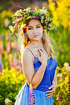 A girl with a flower wreath on her head sits at sunset in a field with purple lupin flowers. Image with selective focus  toning