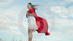 Girl fooling around standing on field in red cloak, playing superhero. cheerful woman plays in red cloak with expression