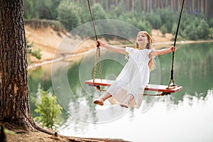 A girl in the forest swinging on a swing. Rope swing on a forest lake. Barefoot girl in a white dress with long hair