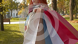 A girl with foxy hair walks through the park with purchases in multi-colored paper bags