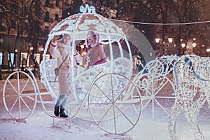 Girl friends taking selfie photo in illuminated fairy tale carriage with horses decorated with lights and garlands for Christmas