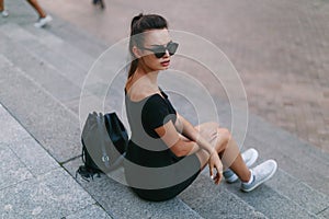 Girl with frown face, sits on the stairs in the city, in a dress with round sunglasses, looking after her shoulder.