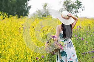 A girl with a full basket of flowers in a light long summer dress stands in the middle of a yellow flower field.