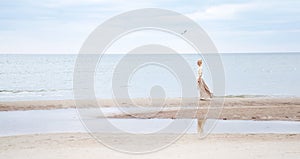 The girl in full growth stands on the beach, ocean. A blond woman in a long beige skirt and a warm white light walks along the coa