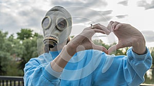 A girl in a gas mask with her hands shows a heart symbol. Alienation in the city. Fashion, ecology and loneliness concepts