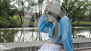 A girl in a gas mask sits on a bench on the river bank. Alienation in the city. Fashion, ecology and loneliness concepts