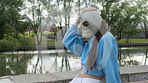 A girl in a gas mask sits on a bench on the river bank. Alienation in the city. Fashion, ecology and loneliness concepts