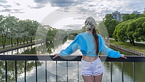 A girl in a gas mask stands on a bridge across the river. Alienation in the city. Fashion, ecology and loneliness concepts