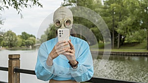 A girl in a gas mask takes a photo on the phone on the river bank. Alienation in the city. Fashion, ecology and loneliness