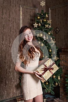 girl genuinely joyfully smiling with a Christmas present in her hands is standing in a gold classic vintage dress