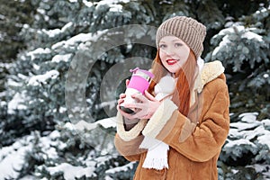 Girl get warm and drink tea in winter park. Fir trees with snow.