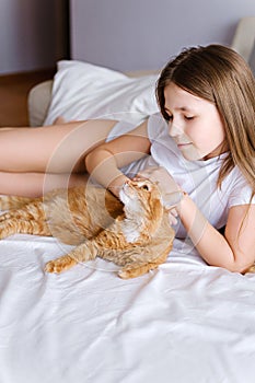 A girl with a ginger cat lies on a white bed. A young girl is stroking a ginger cat while lying on a white sheet