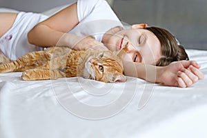 A girl with a ginger cat lies on a white bed. A young girl is stroking a ginger cat while lying on a white sheet