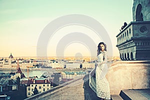 Girl with glamour look. Woman in white wedding dress on city view, fashion. Sensual woman with long hair on balcony