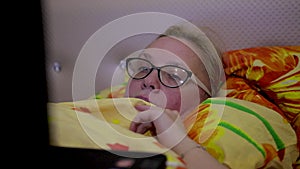 Girl with glasses lying on the bed under a blanket and looking at the laptop screen.