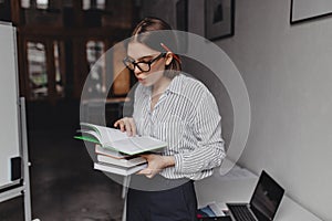 Girl in glasses and office clothes reads books. Snapshot of woman with pencil behind ear on background of workplace with