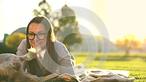 Girl in glasses reading book lying down on a blanket in the park and the small dog runs around and plays around at