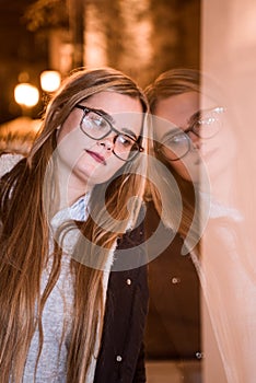 Girl with glasses resting on the glass of a shop, look lost. Sad young girl at night