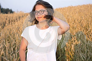 A girl in glasses in a white T-shirt lit by the rays of the setting sun stands in a wheat field and smiles